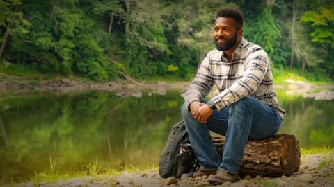 Baratunde, a black man, sits on a log while looking out into nature. The background is a lush, green forest. A calm creek rests behind him.